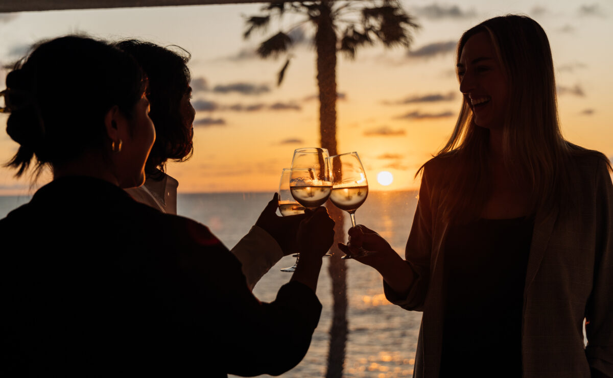 Three people cheers-ing their glasses of wine against a sunset.