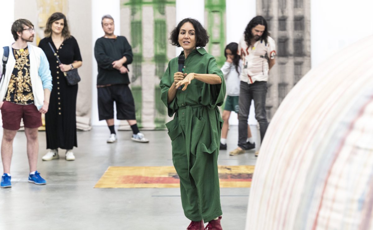 A woman with brown skin and short, curly black hair, wearing a green jumpsuit and red shoes, stands in the middle of artworks with a microphone speaking
