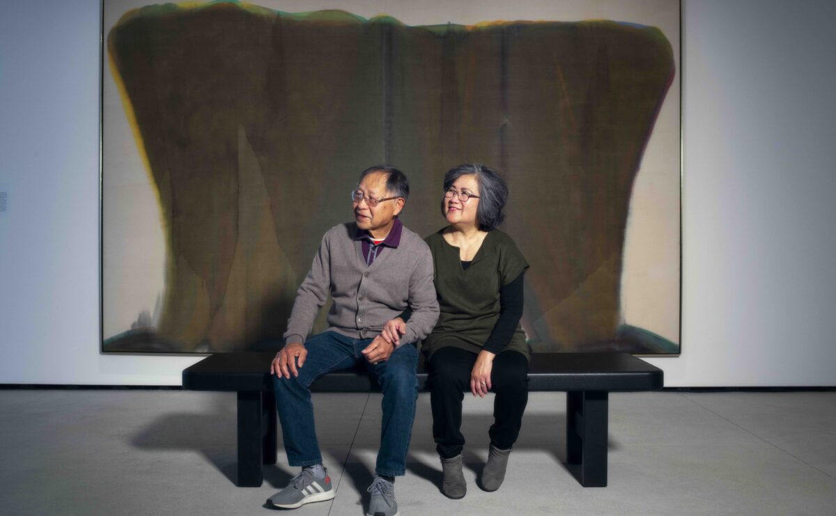 Two people sitting on a black bench inside a gallery looking to their right with a large brown painting behind them