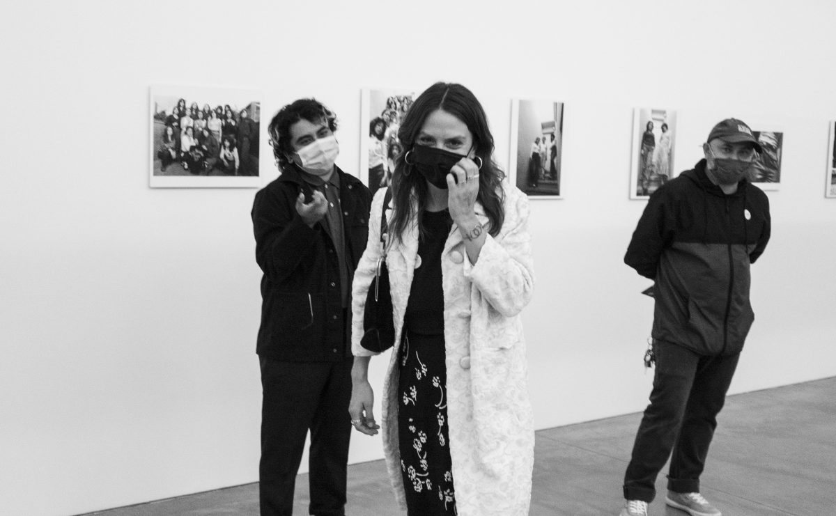 Black and white image of three people standing in a row in front of artwork, all wearing masks