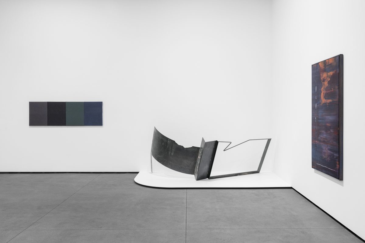 A steel sculpture on the floor flanked by two paintings in a white wall gallery with grey cement floors