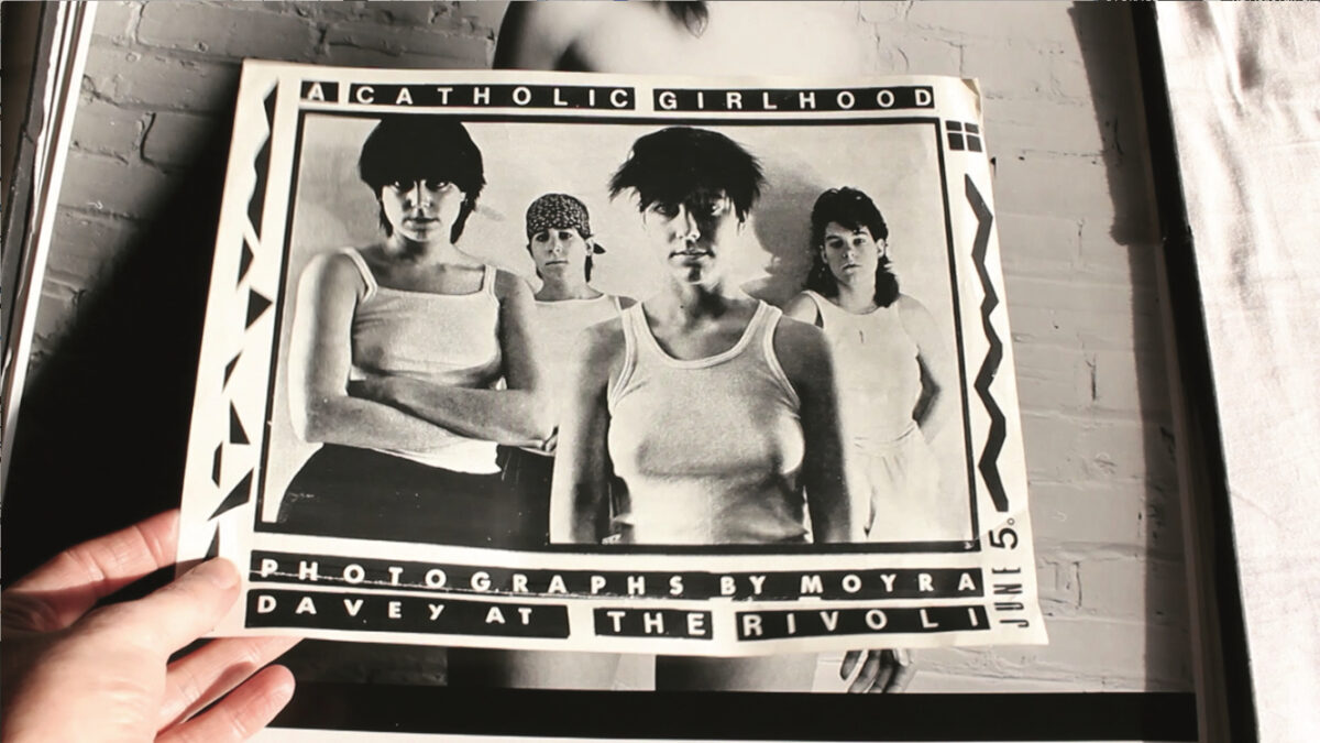 A still from Les Goddesses. A person holds a black and white photograph of four femme-presenting people.