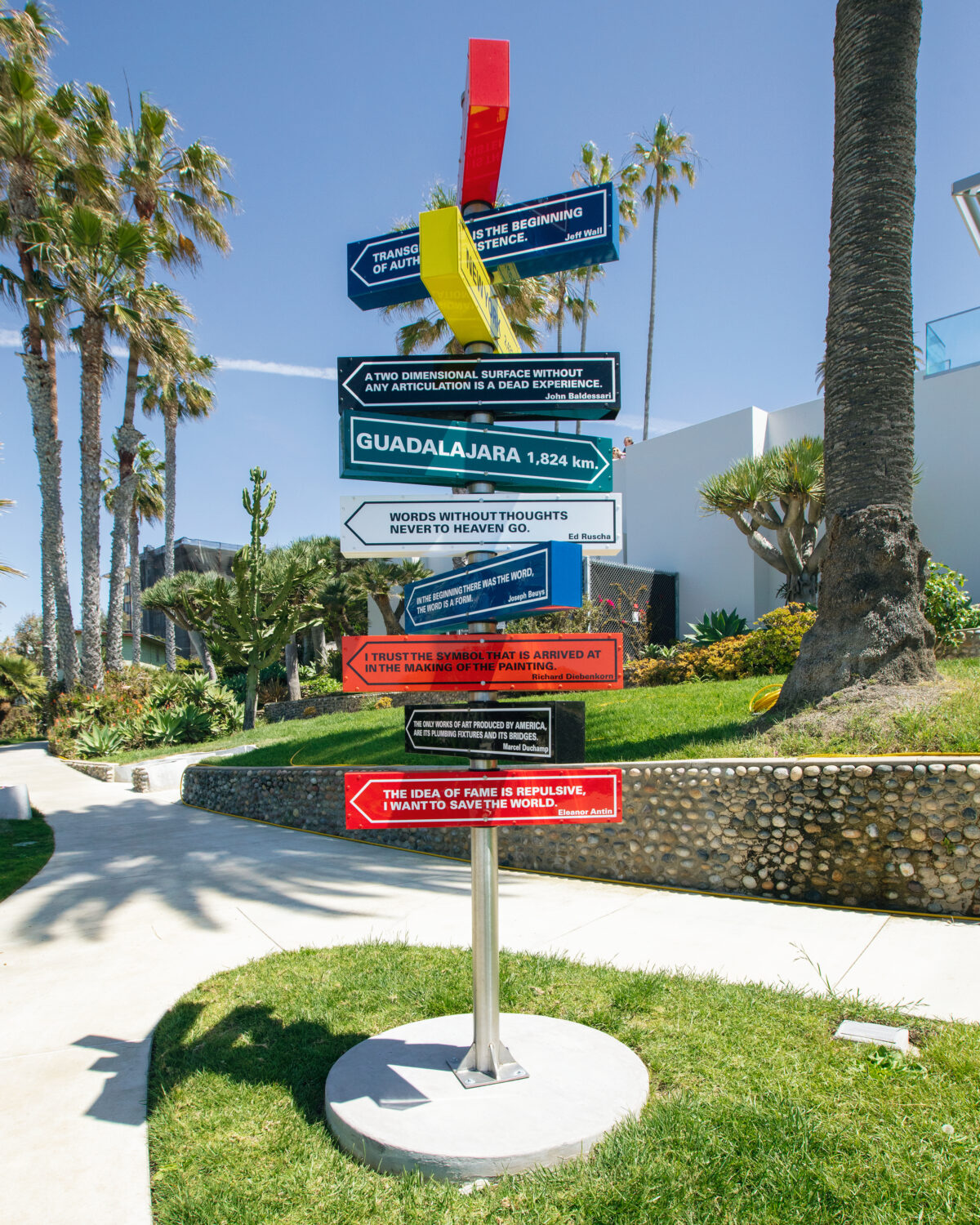 A tall sculpture with multiple, colorful signs pointing in different directions with the name of the city and miles to get there (for example: National City)