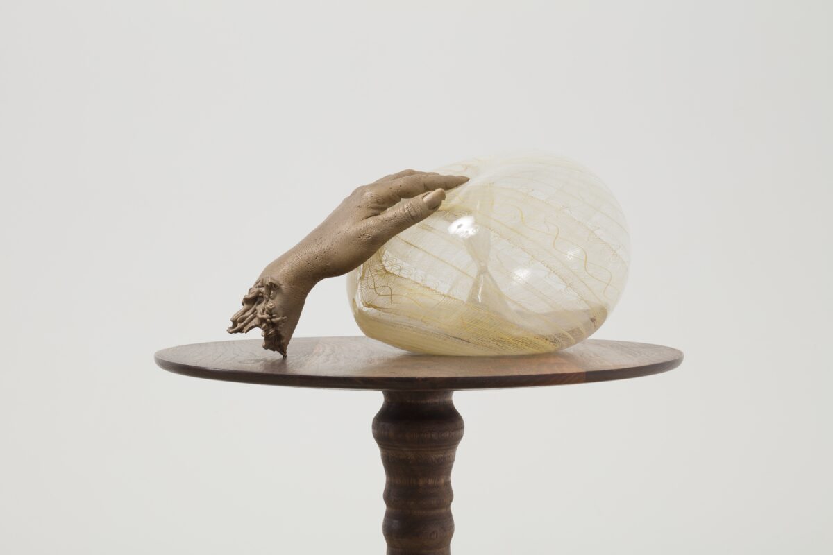 Bronze in the shape of a hand over a hand-blown glass in a circular shape, standing on a walnut pedestal