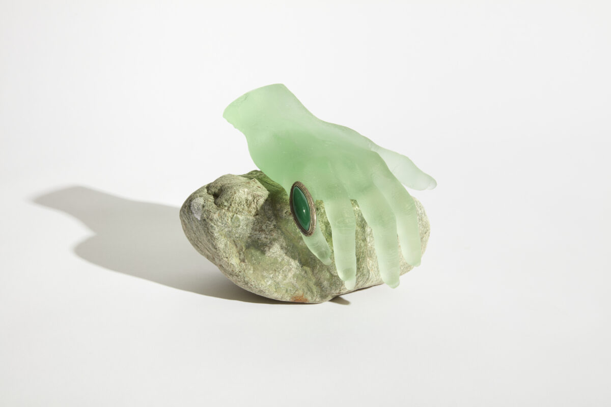 A slightly green cast of a hand with an emerald ring on its pinky. All resting atop a rock.