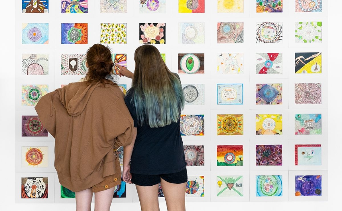 A light skinned teenage girl wearing all brown stands Two teenagers facing a colorful grid of artwork on a white wall