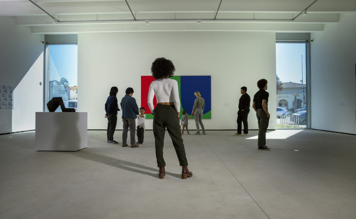 a group of people stand in a gallery observing contemporary art. there is a large colorful painting on the wall, and an abstract metal sculpture on a pedestal.