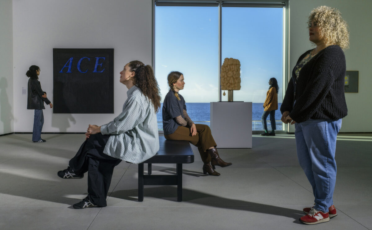 visitors in a gallery look at art on the walls with a view of the ocean in the background