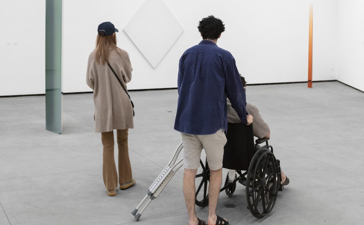 Three people in a white gallery, two standing, one in wheel chair, all looking at three works of light and space