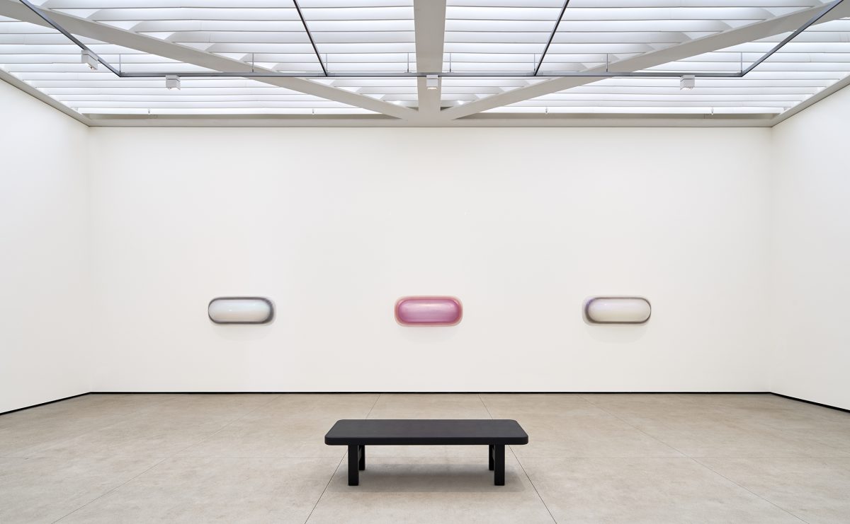 Three bubble-like art pieces on a wall in a white gallery with stark black bench in the foreground