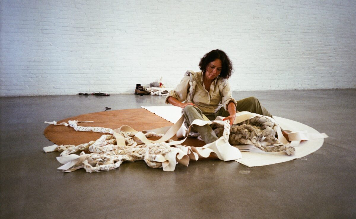 A woman with brown skin sits on the floor while weaving a white circle cloth during a performance piece