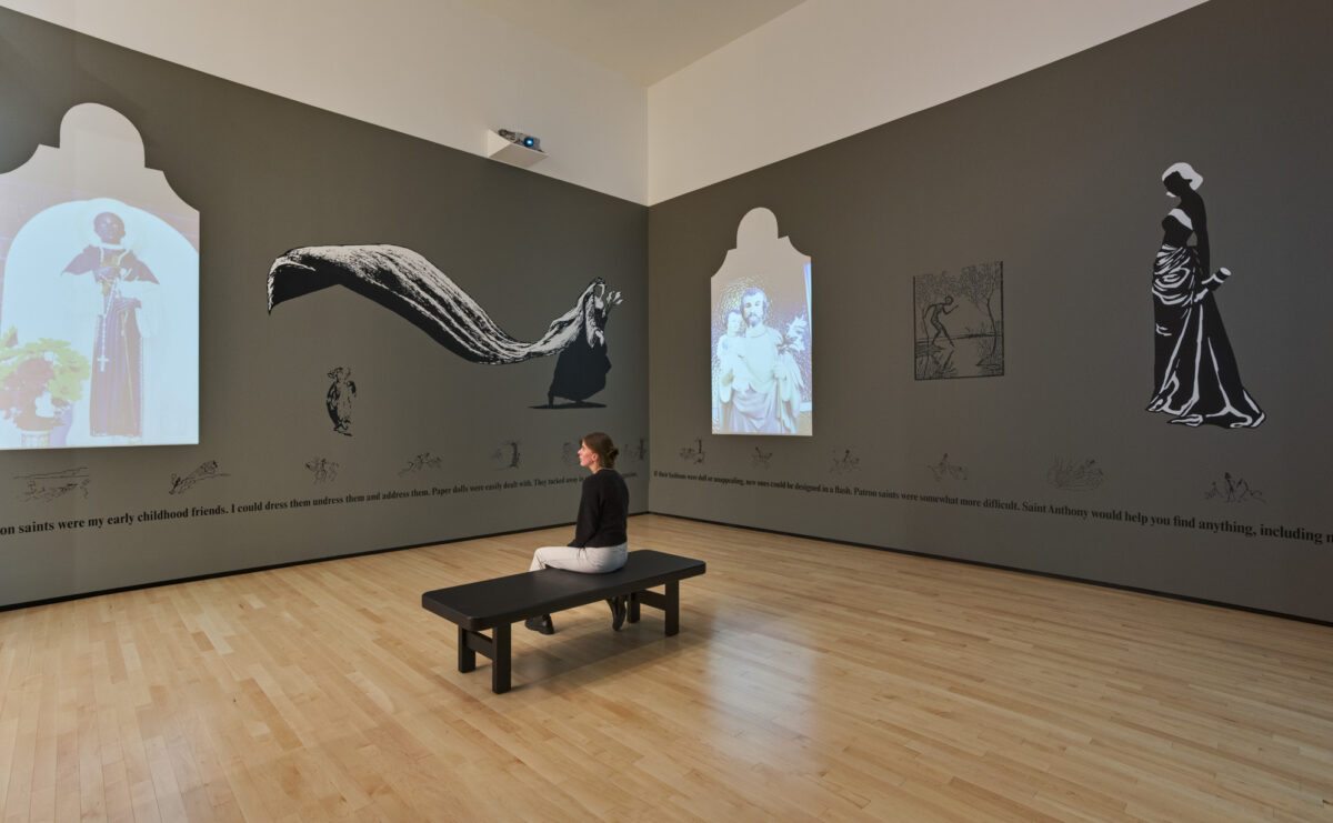 Woman sits on bench in middle of a hard wood gallery, the walls are dark grey with video projections and black and white drawings