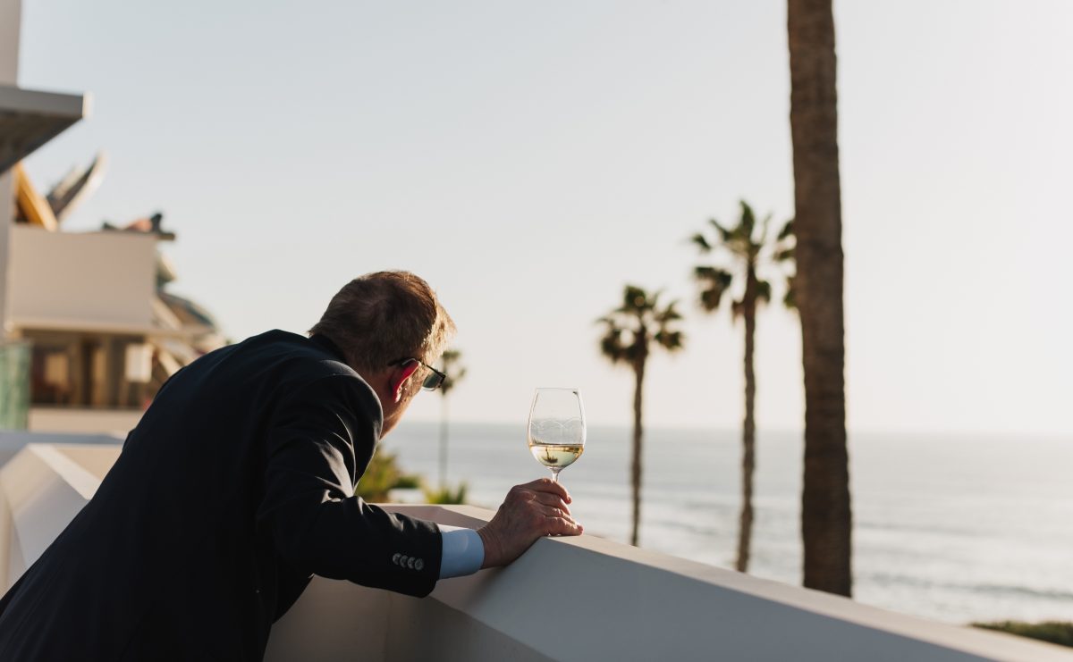 A man in a suit looking out at the ocean over a balcony, a glass of wine in hand