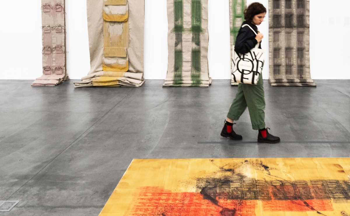 A woman with white skin holding a bag walking between colorful draped fabric on a wall and a colorful paper work on the ground