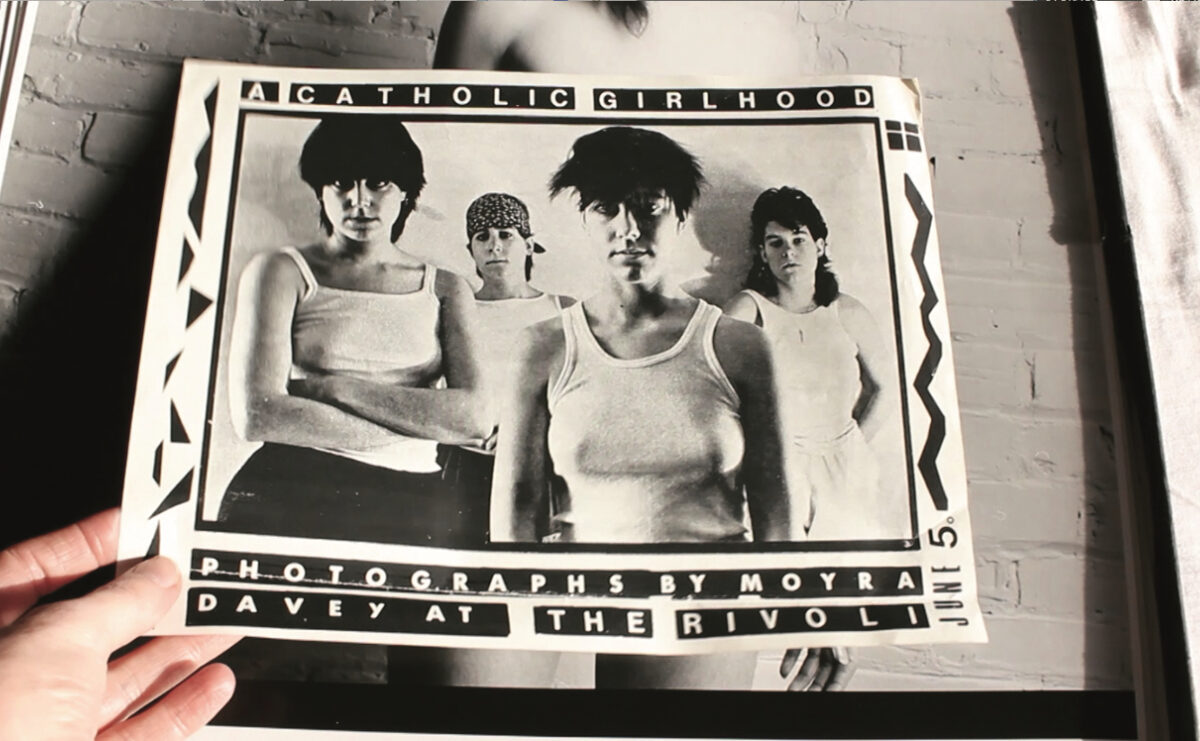 A still from Les Goddesses. A person holds a black and white photograph of four femme-presenting people.