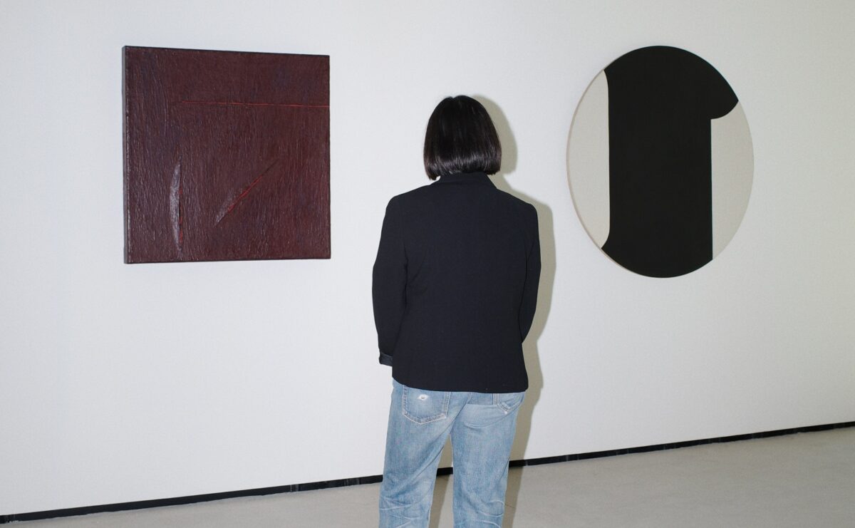 The back of a short woman wearing a dark blazer and light blue jeans looking at minimalist works of art on the wall