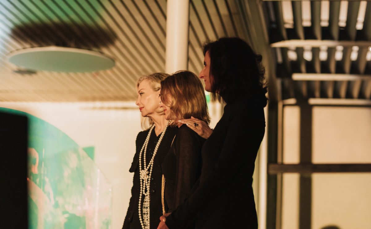 Three woman with light skin wearing all black look out to the left at an event, there are hazy yellow and green colors in the background