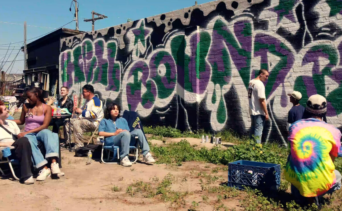 People sit outdoors. Behind them a mural reads "Hell Bound Boys."