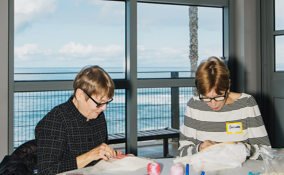Two older adults create art at a table with the ocean in the background.
