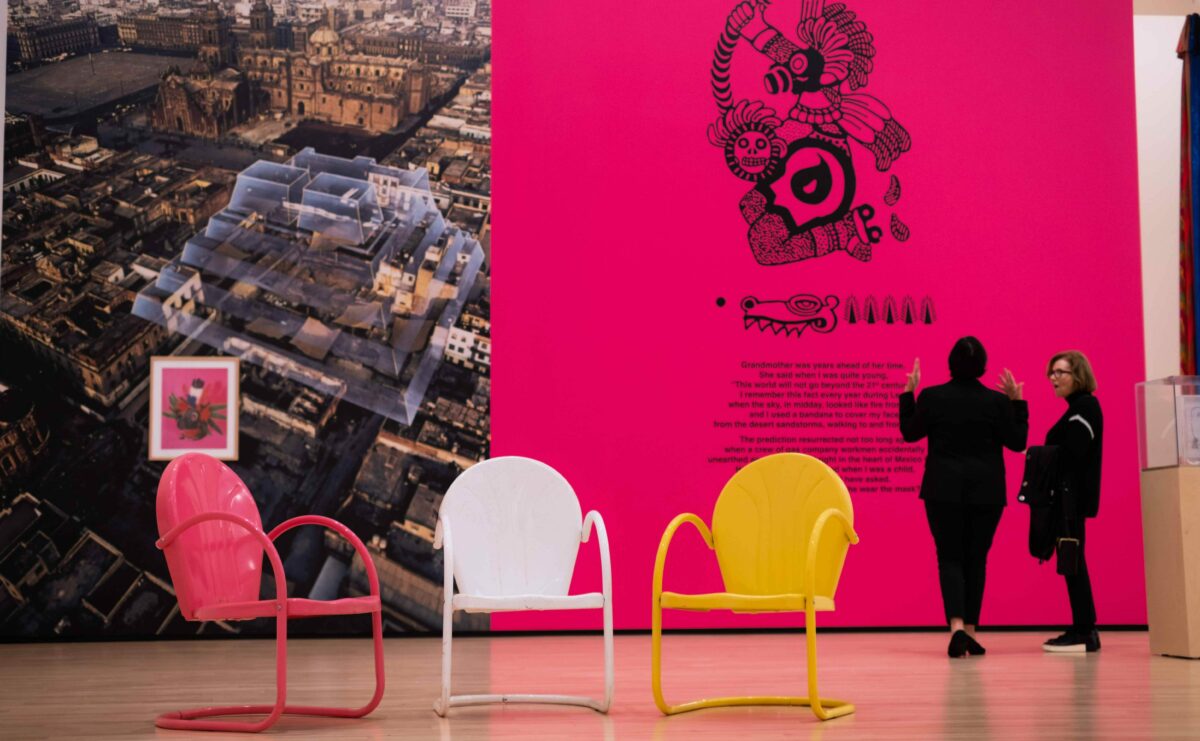 Three brightly colored chairs from Álvarez Muñoz's exhibition in Strauss Galleries. Two people observe her mural in the background.