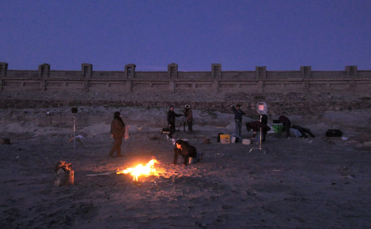 Group of people on the beach near a wall, a campfire in the center, cameras are setup