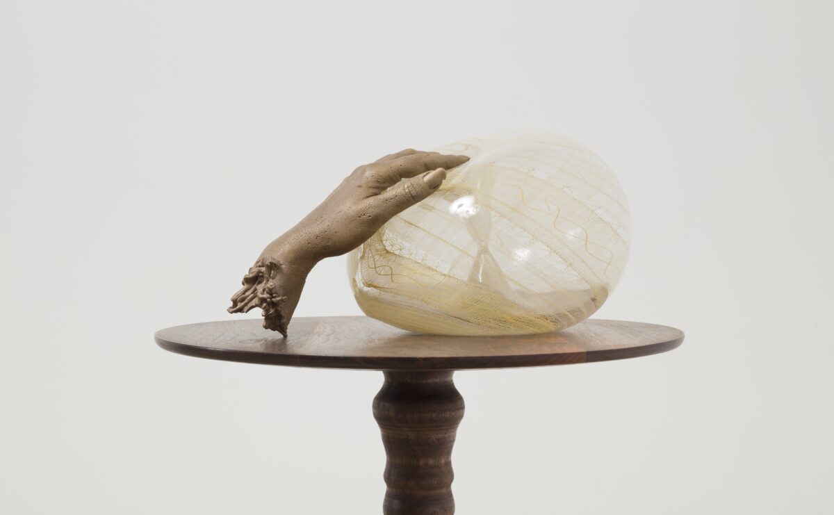 A bronze cast of Akashi's hand delicately on top of a round, yellow, glass blown artwork atop a brown table.