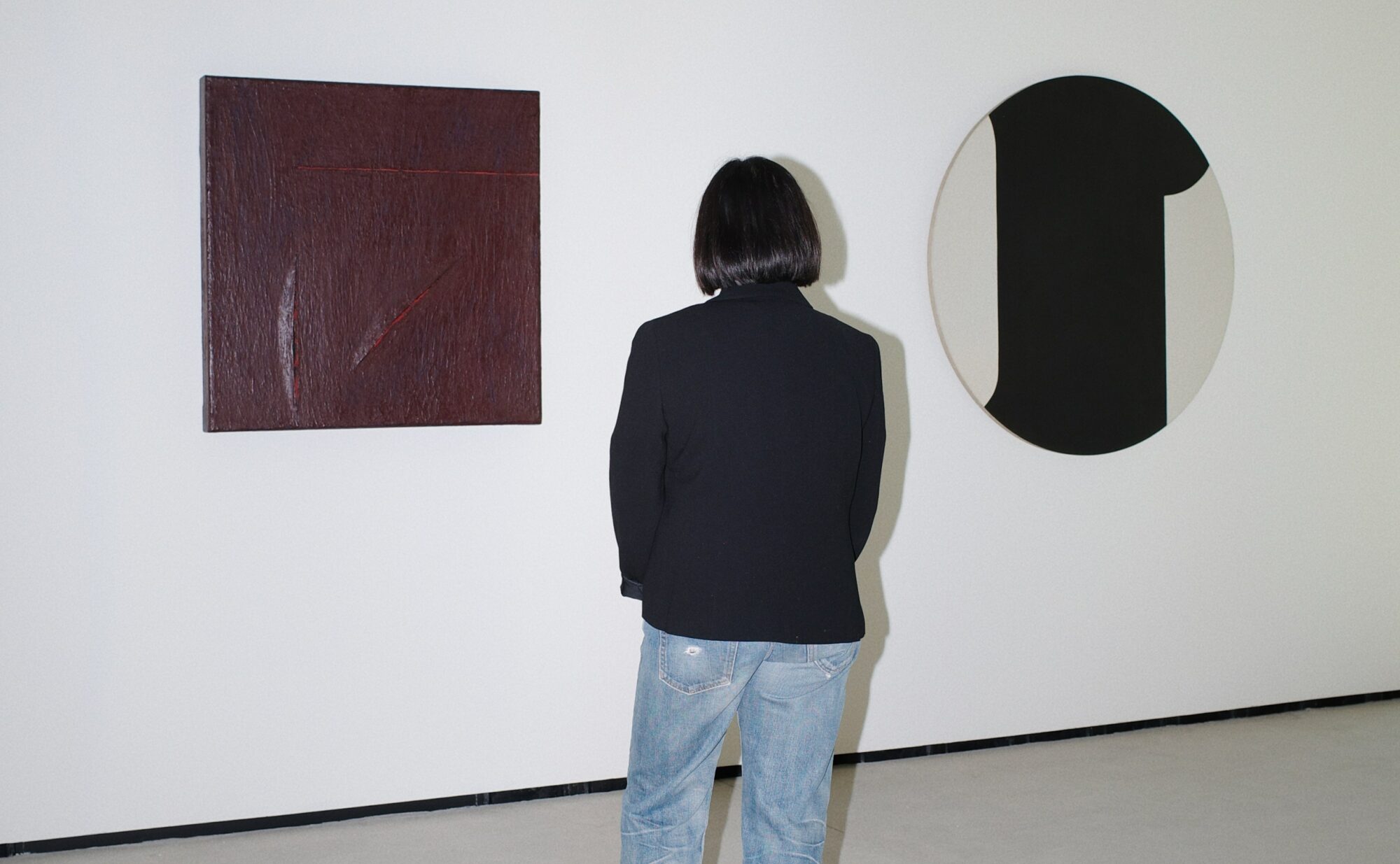 The back of a short woman wearing a dark blazer and light blue jeans looking at minimalist works of art on the wall