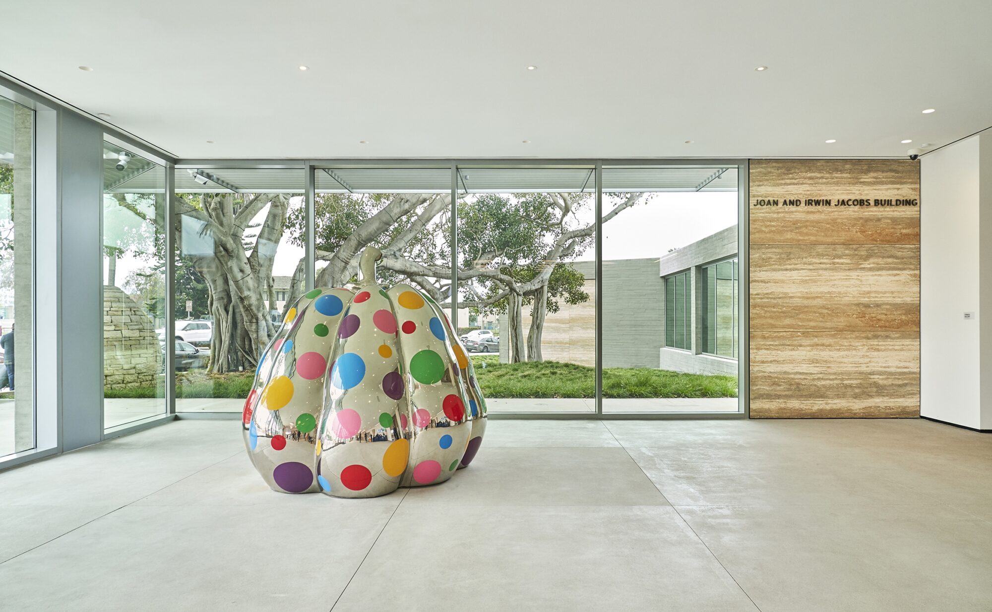 Kusama pumpkin on grey floor with floor to ceiling window backdrop, stone panel on the right that reads "JOAN AND IRWIN JACOBS BUILDING"