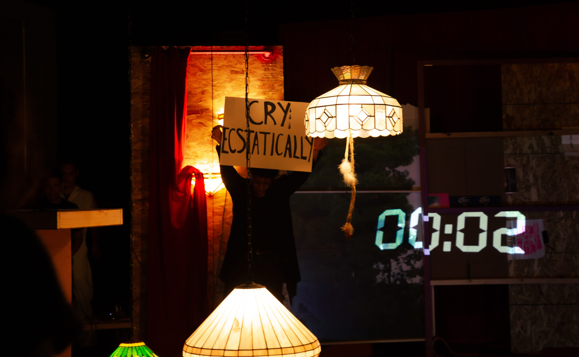 High contrast photo of a living room theatre set. Someone holds up a sign that says cry ecstatically and a clock reads 2 seconds..